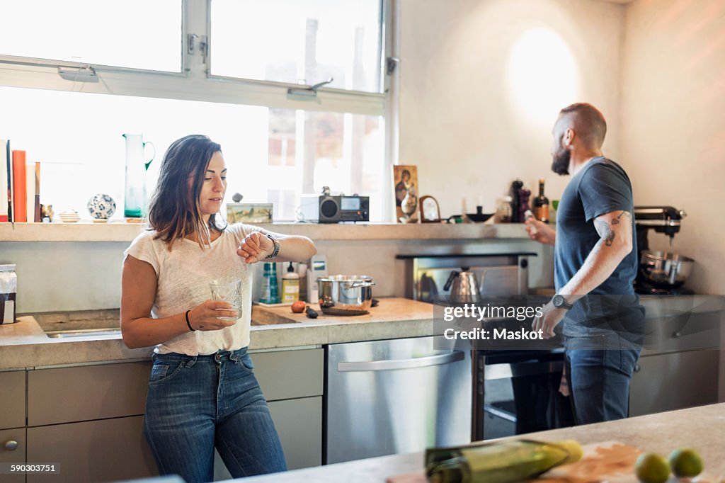 Woman checking time while man looking through window in kitchen