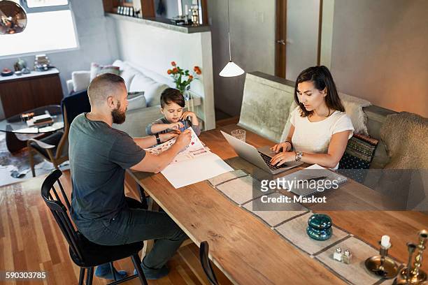 mother working on laptop while father assisting son in doing homework at dining table - homework table stock pictures, royalty-free photos & images