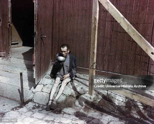 Holocaust survivor drinking from a metal bowl in front of barracks at Buchenwald concentration camp.