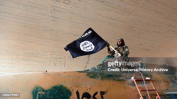 Islamic State fighter waving a flag while standing on captured government fighter jet in Raqqa, Syria, 2015.
