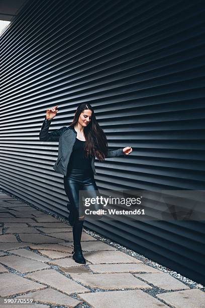 black dressed young woman hopping on one leg in front of black facade - leather pants stockfoto's en -beelden