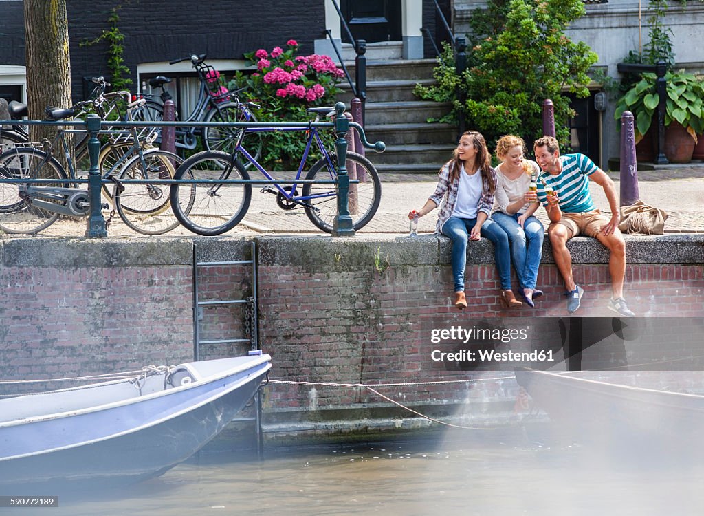 Netherlands, Amsterdam, three happy friends sitting with drinks at town canal