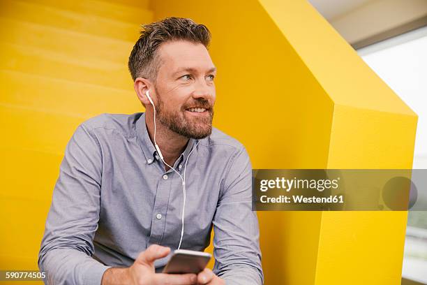 portrait of smiling mature man hearing music with smartphone and earphones sitting on yellow stairs - yellow shirt stock pictures, royalty-free photos & images