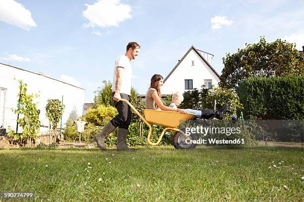 man pushing wheelbarrow with mother and his little son through the garden - wheelbarrow stock-fotos und bilder