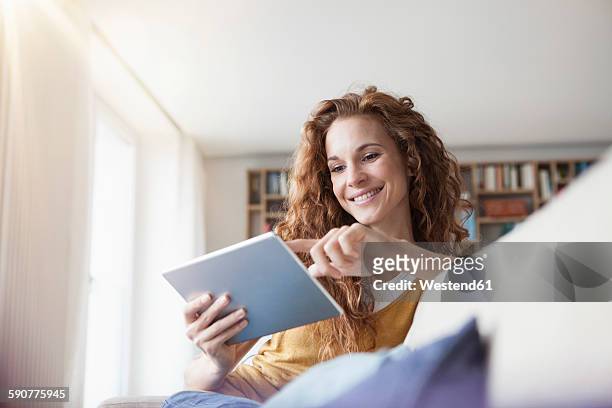 smiling woman at home sitting on couch using digital tablet - usare un tablet foto e immagini stock