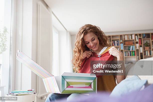 smiling woman at home sitting on couch unpacking parcel with garment - garment tag stock pictures, royalty-free photos & images