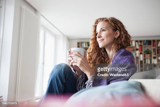 woman at home sitting on couch holding cup - mid adult stock-fotos und bilder