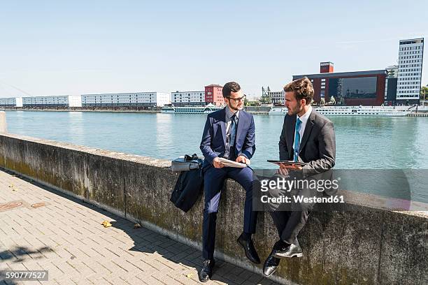 two young businessmen sitting on wall by river, working - mann anzug gebäude objekt draussen stock-fotos und bilder