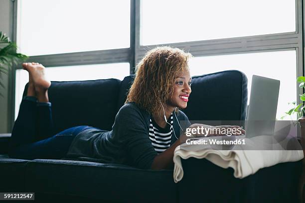 young woman lying on the couch with earphones using laptop - soligt leende bildbanksfoton och bilder
