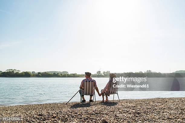 germany, ludwigshafen, back view of senior couple sitting hand in hand on folding chairs at riverside - side by side stock-fotos und bilder