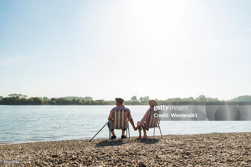 Germany, Ludwigshafen, back view of senior couple sitting hand in hand on folding chairs at riverside