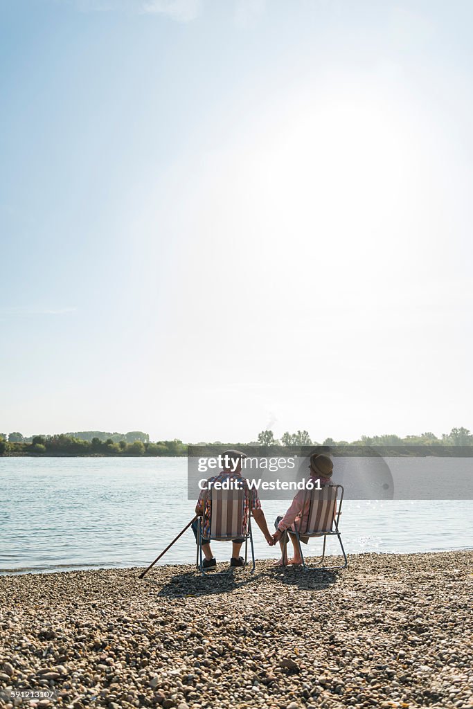 Germany, Ludwigshafen, back view of senior couple sitting hand in hand on folding chairs at riverside