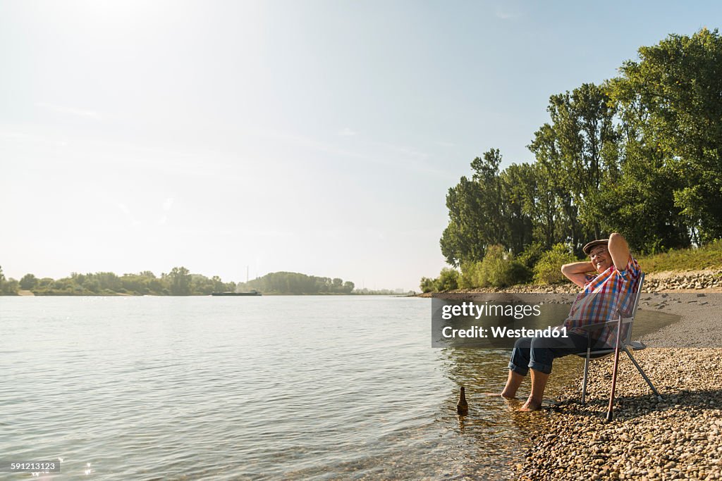 Germany, Ludwigshafen, happy senior man relaxing on folding chair at riverside