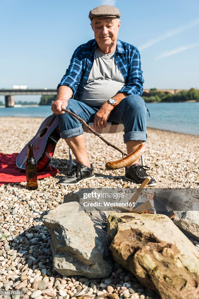 Germany, Ludwigshafen, portrait of smiling senior man barbecueing sausage on the beach