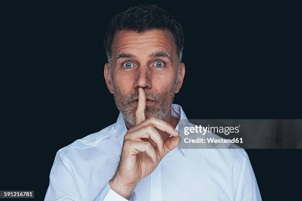 portrait of man with finger on his mouth in front of black background - silêncio - fotografias e filmes do acervo