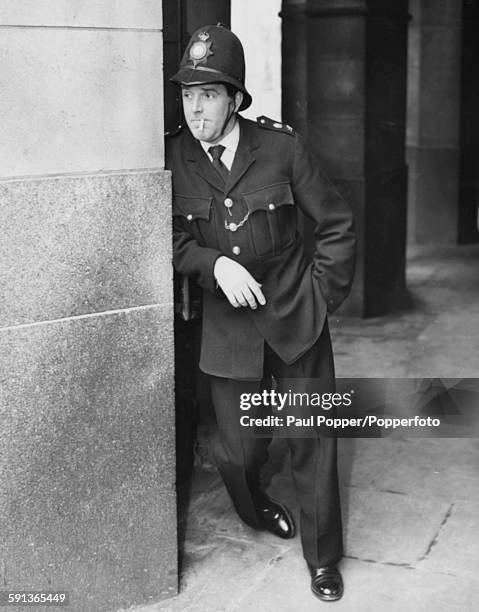 English actor Brian Rix wearing police uniform playing the character of PC Percy Hobson, smoking a cigarette during rehearsals for the BBC show 'A...