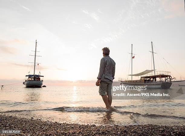 man stands on beach, looks past boats at sunrise - stadt antalya stock-fotos und bilder