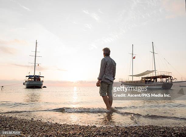 man stands on beach, looks past boats at sunrise - antalya stock pictures, royalty-free photos & images