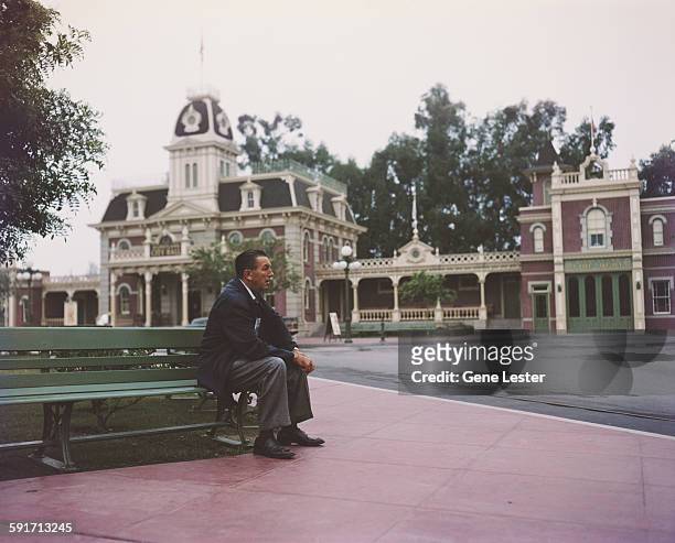 Portrait of American movie producer, artist, and animator Walt Disney as he sits on a bench in his Disneyland amusement park, Anaheim, California,...