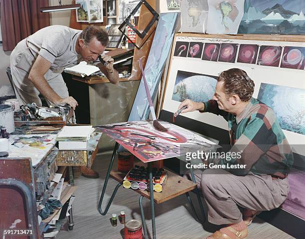 American movie producer, artist, and animator Walt Disney looks through a lens at an oversized paintbrush while an unidentified man drips paint from...