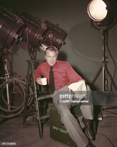Backstage portrait of American actor Gary Cooper as he sits on an equipment box and leans back against a stepladder, mid 1950s. He wears a red oxford...