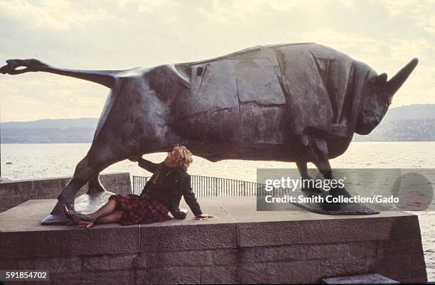 Mature female tourist jokingly pretends to grab the crotch area of a large metal statue of a bull, 1975. .