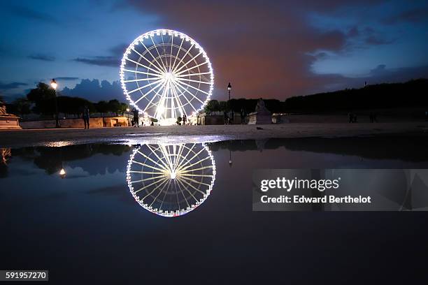 The ferris wheel "Roue de Paris" reflects into a water puddle on a rainy summer day, during night time, in the garden "Jardin des Tuileries", near...