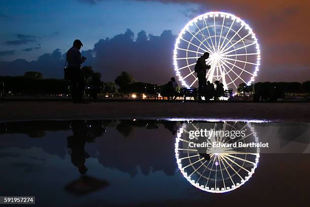The ferris wheel "Roue de Paris" reflects into a water puddle on a rainy summer day, during night time, in the garden "Jardin des Tuileries", near...