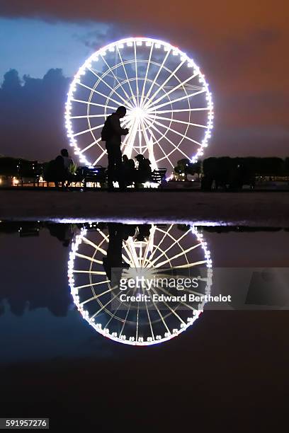 The ferris wheel "Roue de Paris" reflects into a water puddle on a rainy summer day, during night time, in the garden "Jardin des Tuileries", near...