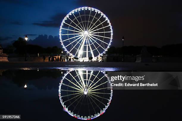 The ferris wheel "Roue de Paris" reflects into a water puddle on a rainy summer day, during night time, in the garden "Jardin des Tuileries", near...