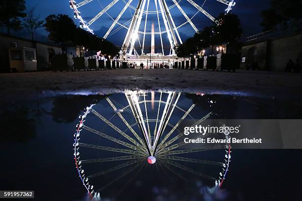 The ferris wheel "Roue de Paris" reflects into a water puddle on a rainy summer day, during night time, in the garden "Jardin des Tuileries", near...