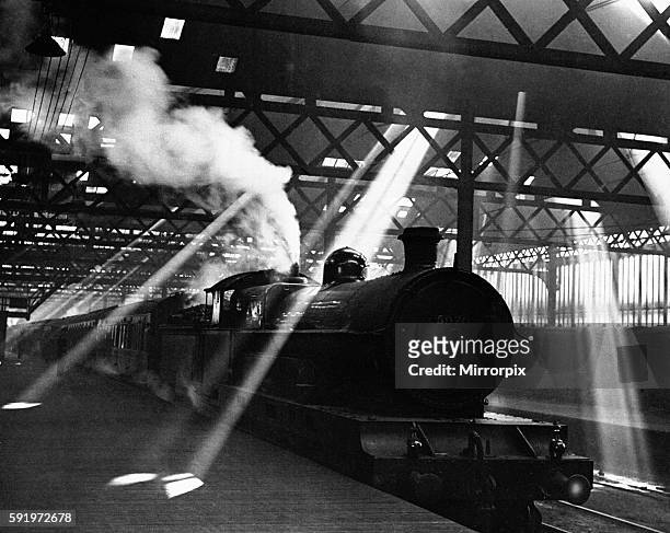 Through the murk and lattice work roof, a pyramid of light shafts surround a London & North Western Railway Claughton class 4-6-0 No. 5970 Patience...
