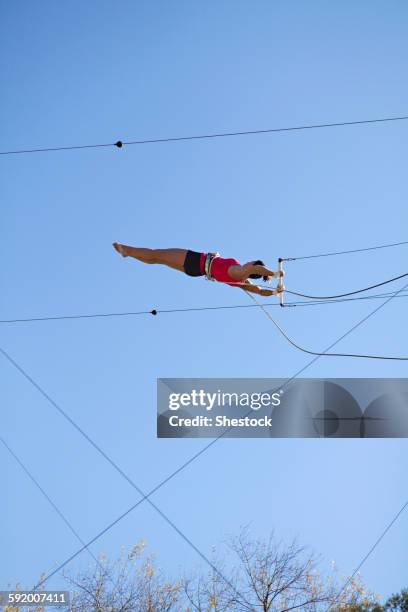 acrobat hanging from trapeze under blue sky - drahtseilakt stock-fotos und bilder
