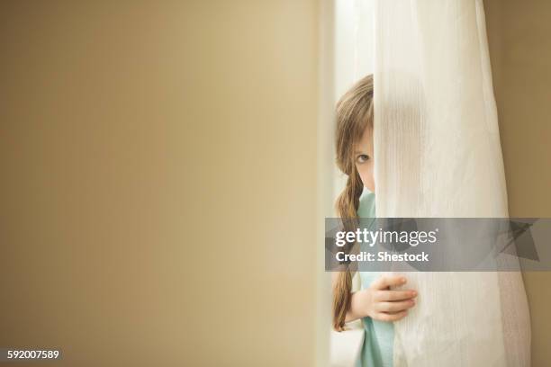 shy caucasian girl peeking around curtain - shy foto e immagini stock