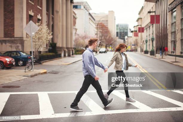 caucasian couple holding hands crossing city intersection - man side way looking stock pictures, royalty-free photos & images