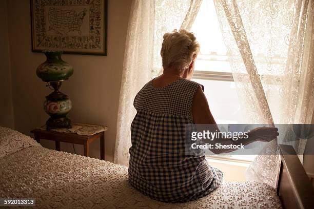 pensive older woman looking out bedroom window - old bed stockfoto's en -beelden