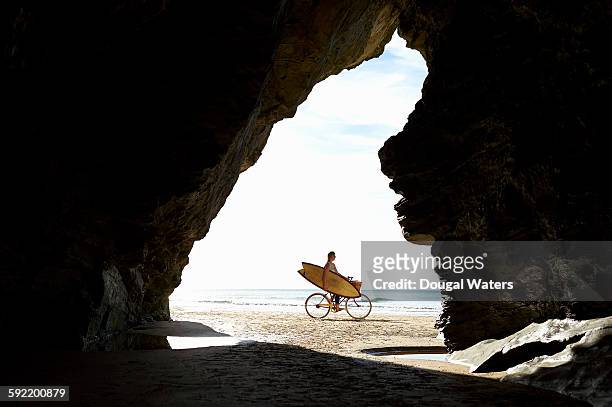 woman cycling on beach holding surfboard. - beach surfer stock-fotos und bilder
