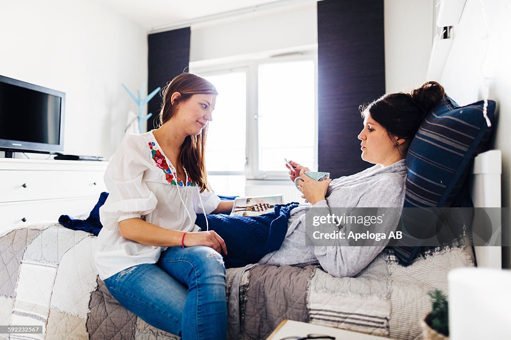 Woman looking at ill female friend holding medicine on bed
