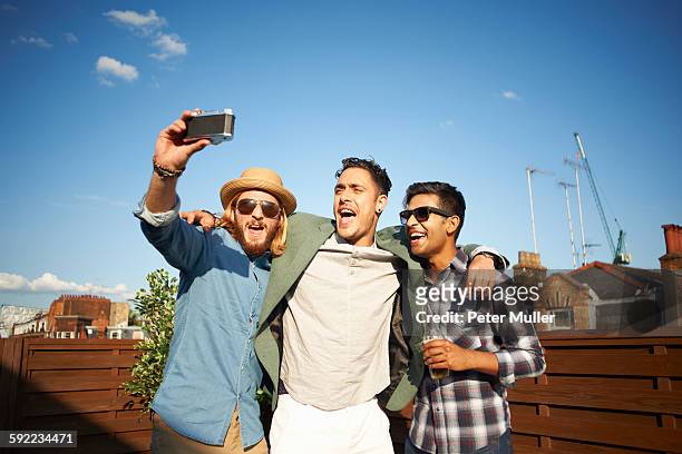 three male friends taking camera selfie at rooftop party - tres amigos fotografías e imágenes de stock