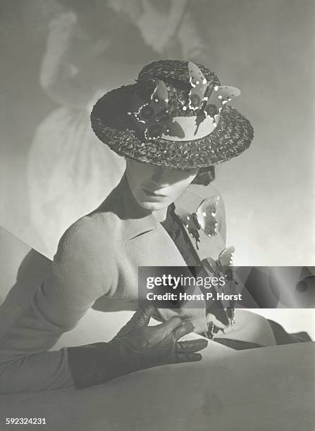 Model, posed with her head down, in a collarless jacket with large button holes and large butterflies on the edges, and matching hat with tow more...