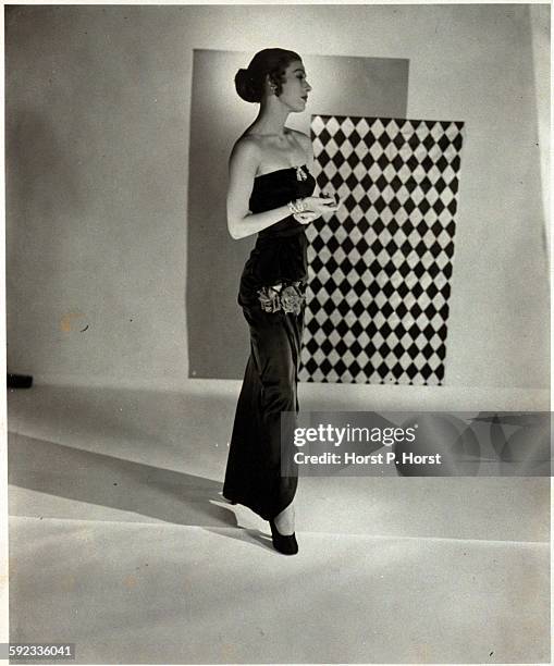 Model standing and wearing a bare-shouldered, ankle-length dress in black rayon satin with two roses at the thigh, by Adele Simpson, and diamond...