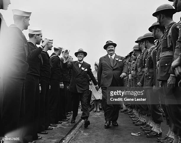Comedy duo Stan Laurel and Oliver Hardy laugh and smiles as they walk along a line of soldiers and sailors, late 1930s or early 1940s.