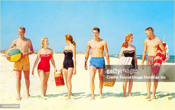Vintage photograph of three young couples wearing bathing suits, carrying picnic supplies and a beach ball, in the sand at an ocean beach, 1950s.