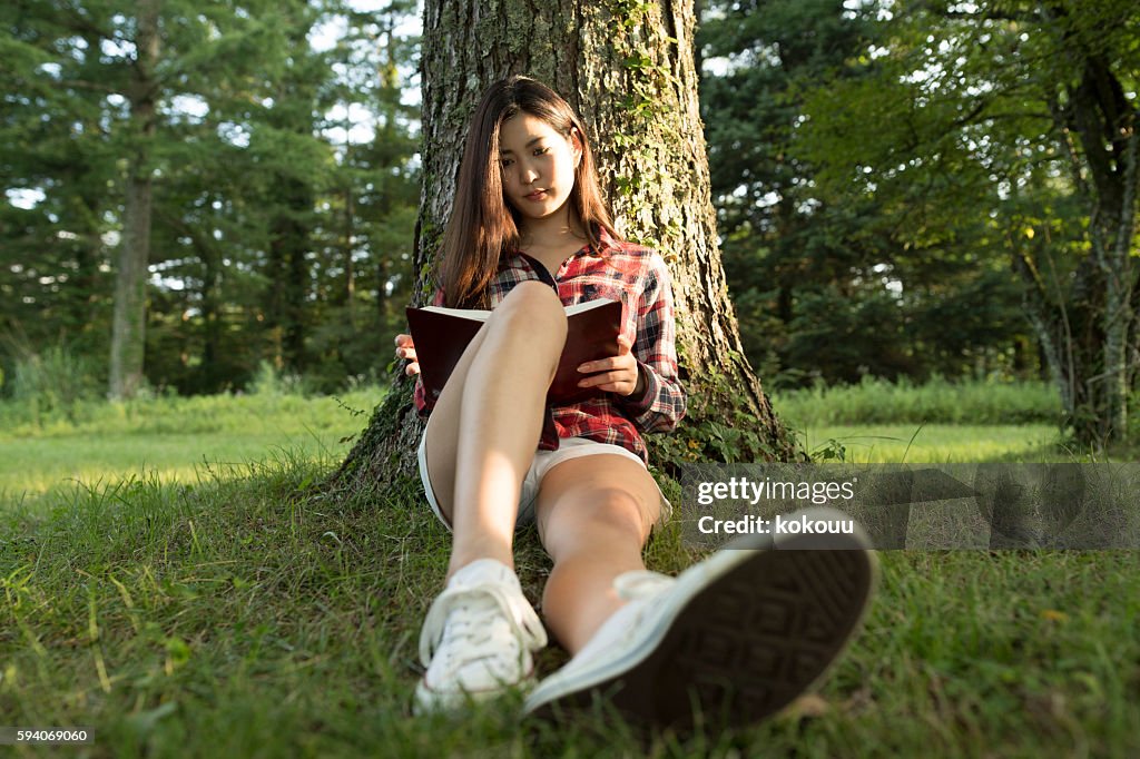 Girl reading a book leaning on a tree.