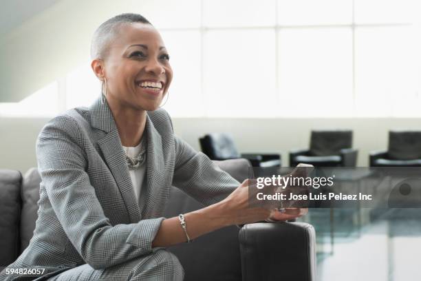 black businesswoman using cell phone on sofa in office - black suit stock-fotos und bilder