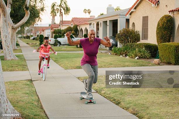 mother and daughter riding skateboard and bicycle on sidewalk - skaten familie stock-fotos und bilder