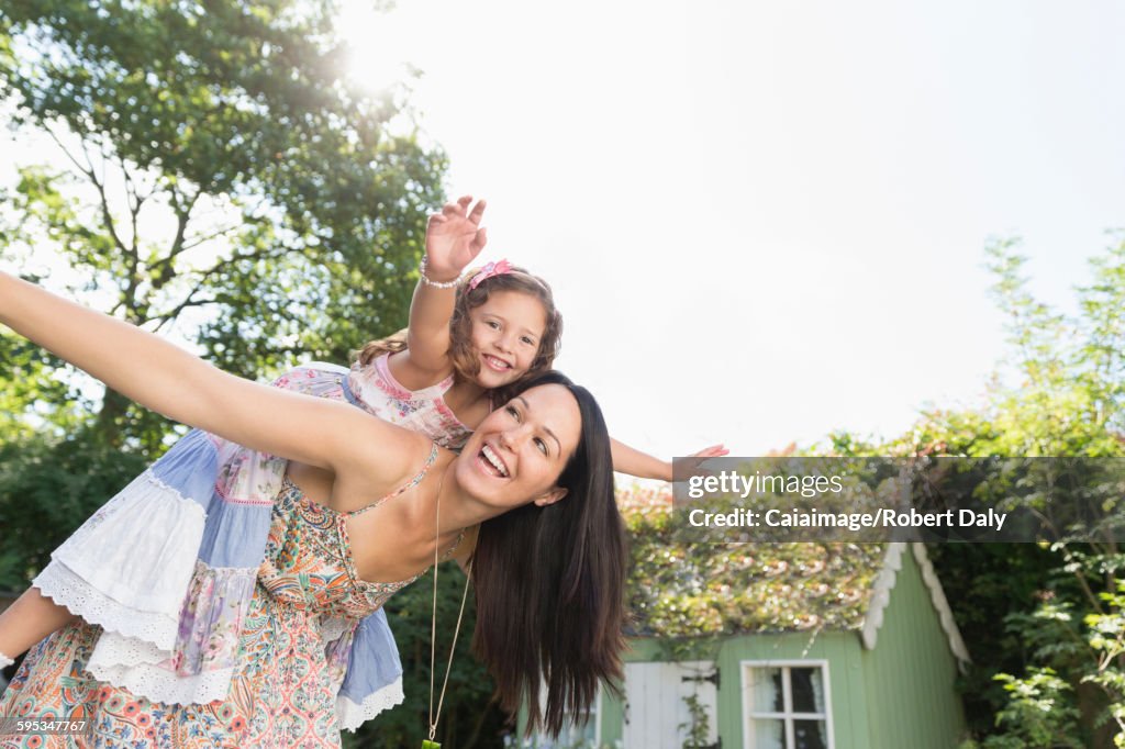 Carefree mother piggybacking daughter with arms outstretched in backyard