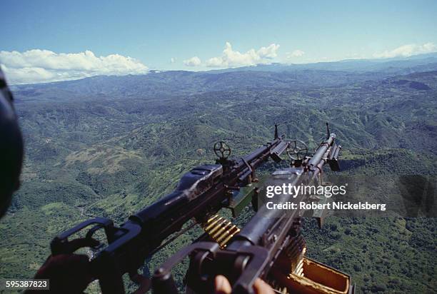 View, along the barrels of a pair of helicopter-mounted, M-60 machine guns, over rural terrain, Morazan department, El Salvador, October 23, 1984....