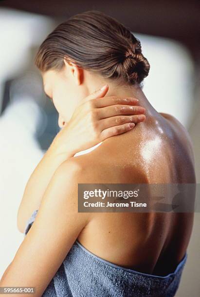 young woman wrapped in towel rubbing cream on neck, rear view - huile de massage photos et images de collection