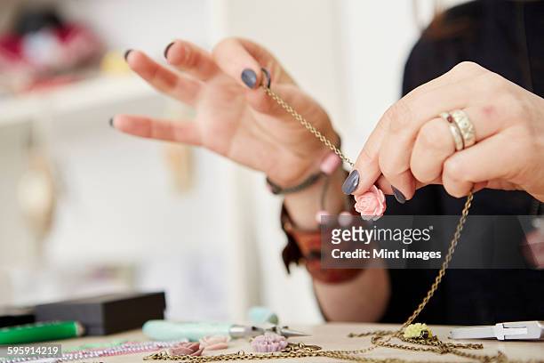 a woman seated at a workbench holding a gold chain with a small floral pendant, making jewellery.  - jeweller stock pictures, royalty-free photos & images